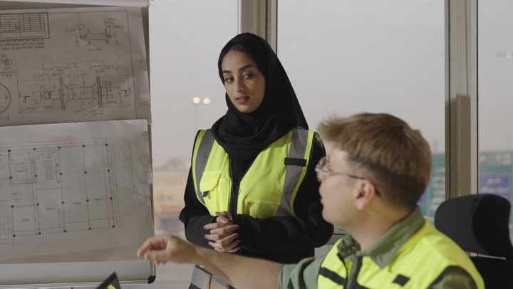 الاستماع لكافة الاراء بين اعضاء الفريق الواحد , رجل اجنبي يرتدي الزي الكاجوال و نظارات يتناقش مع الزميلة داخل الاجتماع , مهندسة عربية خليجية سعودية محجبة ترتدي العباءة السوداء توضح الرسومات الهندسية المعلقة لباقي اعضاء الفريق