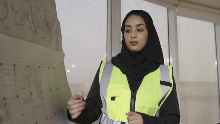 مهندسة عربية خليجية سعودية محجبة ترتدي العباءة السوداء توضح لباقي اعضاء فريق العمل المخططات الهندسية المعروضة , الاجتماع المعقود داخل مقر الشركة