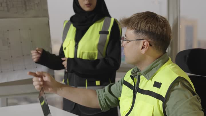 تقبل الاراء المختلفة بين الفريق , امرأة عربية خليجية سعودية محجبة تشرح الرسومات الهندسية المعروضة , مهندس اجنبي يرتدي الزي الكاجوال و السترة الواقية يتحاور مع زميلة العمل حول المخططات , اجتماع في مقر الشركة , مفهوم الديمقراطية