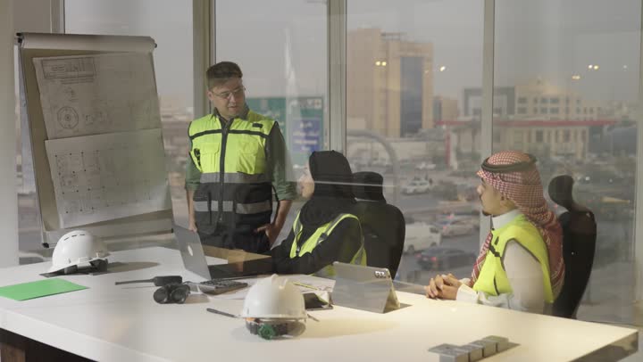 A Saudi Arabian Gulf Arab female engineer wearing a black abaya points with her index finger at the architectural drawings, a foreign man in a safety vest explains the hanging engineering plans, and a Saudi Arabian Gulf Arab man in a white thobe and a colored ghutrah attends a meeting with team members, teamwork.