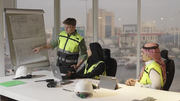 رجل اجنبي يرتدي الزي الكاجوال و السترة الواقية يقف بجانب المخططات الهندسية المعلقة , امرأة عربية خليجية محجبة ترتدي العباءة السوداء تجلس مع فريق العمل في الاجنماع , رجل عربي خليجي سعودي يرتدي الثوب الابيض و الشماغ الملون يشير باصبع السبابة الي شئ ما في الرسومات المعروضة , الادوات الهندسية على الطاولة , الحفاظ على تعليمات السلامة و الامان