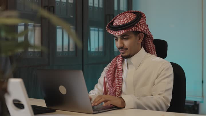 Expressions of smiling during working hours, a Saudi Gulf Arab man wearing a colored ghutrah and a white thobe sitting inside the office, writing and taking notes on the computer, relying on modern advanced technological devices to perform tasks.