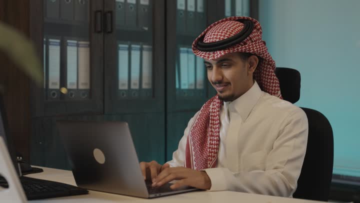 القيام بالمهمات اليومية الروتينية بسهولة على جهاز الحاسوب , رجل عربي خليجي سعودي يرتدي الثوب الابيض و الشماغ الاحمر يجلس امام المكتب , ايماءات السعادة مع الجدية اثناء ساعات العمل