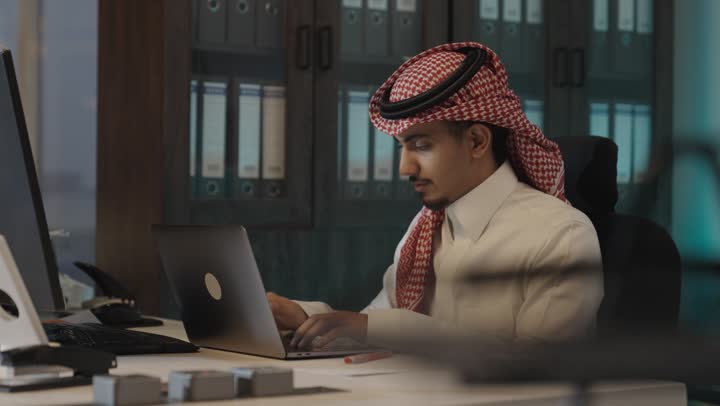 The integration of traditional old methods and modern advanced methods within companies, an Arab Gulf Saudi man wearing a white thobe and a colored ghutrah is taking notes with a ballpoint pen, sitting in front of a portable computer.