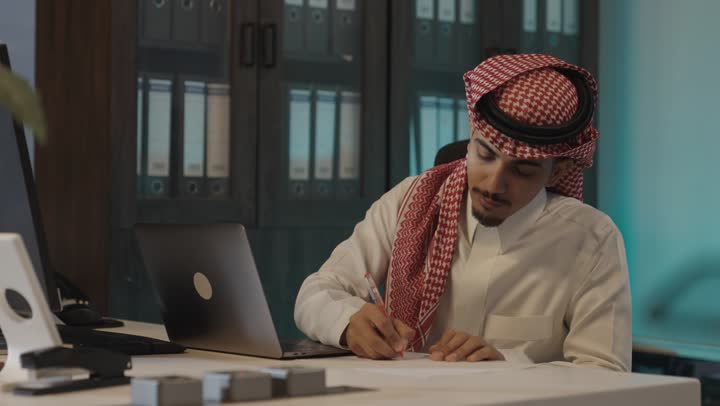 نقل المعلومات من جهاز الحاسب الي الورقة , رجل عربي خليجي سعودي يرتدي الثوب الابيض و الشماغ الملون يكتب القارير الخاصة بالاعمال اليومية على المكتب , غرفة الارشيف داخل الشركةلحفظ المجلدات القديمة