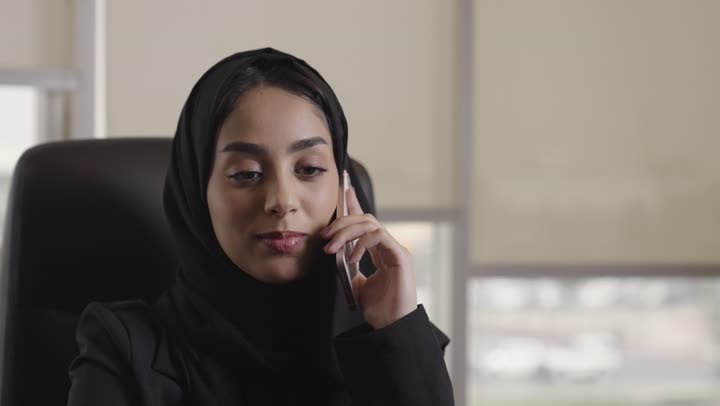 امرأة عربية خليجية سعودية محجبة ترتدي الزي الرسمي تعمل في شركة , اجراء مكالمات هاتفية خاصة بالعمل , استخدام اجهزة تقنية حديثة متعددة , الجهاز اللوحي على المكتب