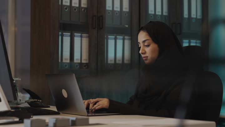 الاجهزة التقنية الحديثة تؤدي الاعمال بدقة و تنظيم , امرأة عربية خليجية سعودية محجبة ترتدي العباءة السوداء تجلس امام جهاز الحاسب المتنقل , تخزين المعلومات بسهولة , ايماءات الاحترافية و الجدية في العمل
