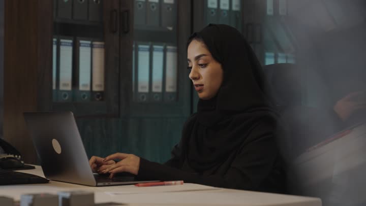الكتابة على الحاسب المتنقل , امرأة عربية خليجية سعودية محجبة ترتدي العباءة السوداء تمارس الاعمال الكتابية بين الحاسوب و الاوراق و القلم , غرفة تخزين المجلدات الكبيرة الارشيفية