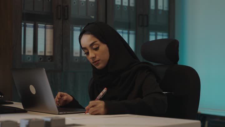 العمل المنظم بالدمج بين الاساليب الحديثة و القديمة , امرأة عربية خليجية سعودية محجبة ترتدي العباءة السوداء تدون الملاحظات من الحاسب على الاوراق , الجلوس داخل غرفة الارشيف