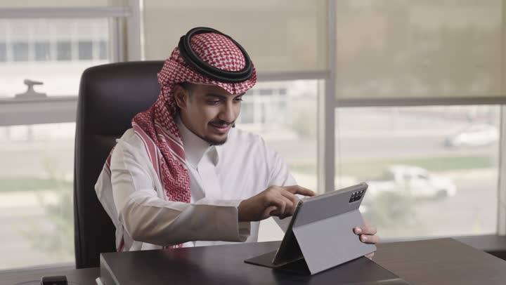 استخدام خاصية تقريب الصور لرؤية اوضح ,رجل عربي خليجي سعودي يرتدي الشماغ الملون الاحمر و الثوب الابيض يستخدم الجهاز اللوحي للقيام بالاعمال المطلوبة , الجلوس داخل مقر الشركة
