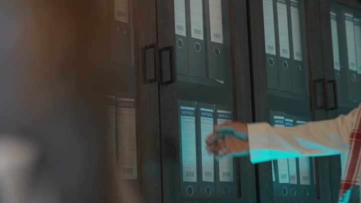 استخدام الطرق القديمة في حفظ البيانات الخاصة بالعمل , رجل عرب خليجي سعودي يرتدي الثوب الابيض و الشماغ الملون الاحمر يلتقط المجلد الورقي الكبير , الوقوف امام دولاب الحفظ