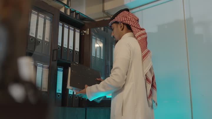 النظر بايماءات السعادة و الابتسامة الى الاوراق المحفوظة , رجل عربي خليجي سعودي يرتدي الثوب الابيض و الشماغ الاحمر الملون , الوقوف داخل غرفة الارشيف , حفظ البيانات بالطرق القديمة
