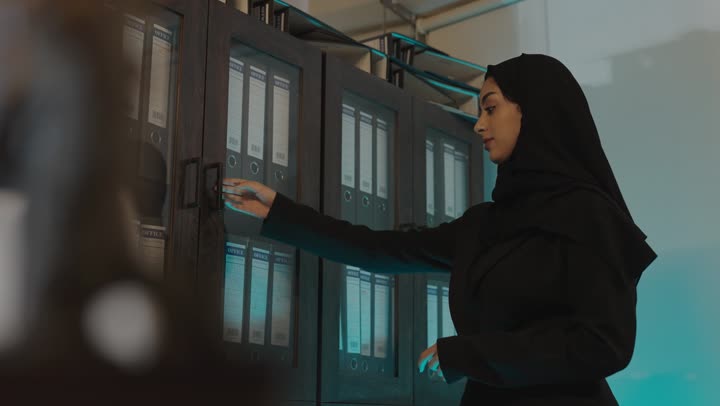 فتح دولاب المجلدات القديمة , امرأة عربية خليجية سعودية محجبة ترتدي العباءة السوداء تحمل الاوراق المحفوظة في غرفة الارشيف , البحث عن المخطوطات القديمة