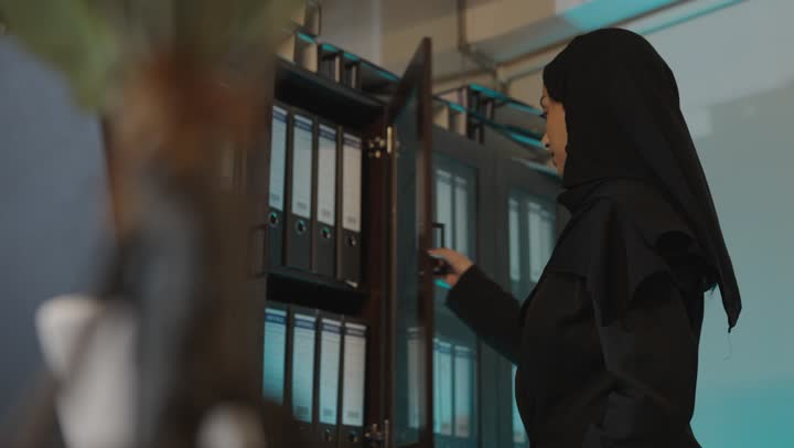 الوقوف امام دولاب حفظ المجلدات الورقية , امرأة عربية خليجية سعودية ترتدي العباءة السوداء تحمل المجلد الكبير , عملية البحث عن البيانات القديمة