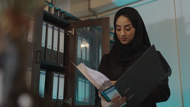 الاحتفاظ بالمعلومات في المجلدات الورقية , امرأة عربية خليجية سعودية محجبة ترتدي العباءة السوداء تبحث عن شئ ما في الاوراق المحفوظة في الارشيف , ايماءات التركيز و الجدية