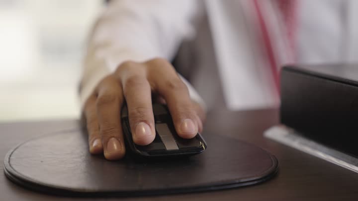 رجل عربي خليجي سعودي يرتدي الثوب الابيض و الشماغ الملون يمسك فأرة الحاسوب للوصول للمجلدات الخاصة بالعمل , قطعة جلدية ناعمة سوداء على المكتب لتحريك جهاز التأشير بسلاسة , تصوير مقرب