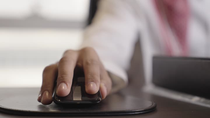 قطعة جلدية سوداء ناعمة لتحريك جهاز التأشير بسهولة , رجل عربي خليجي سعودي يرتدي الثوب الابيض ينقر على فأرة الحاسوب للوصول الي المعلومات المحفوظة ,