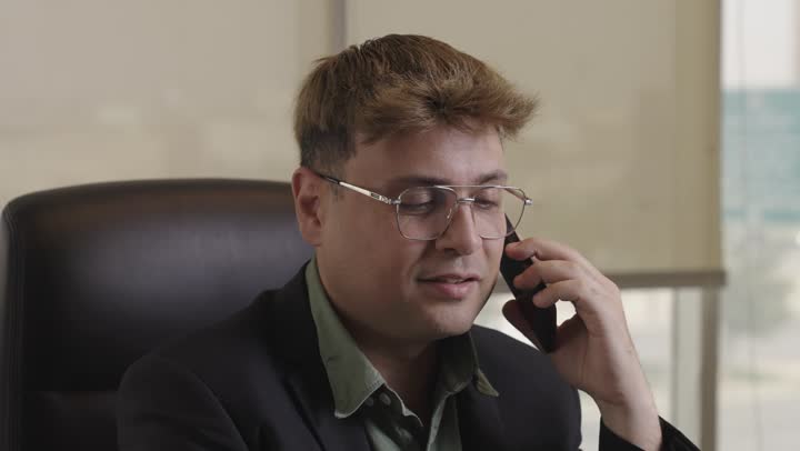 تعابير القبول و اعطاء الموافقة اثناء المحادثة الهاتفية داخل مقر الشركة , رجل اجنبي يرتدي النظارة و البذلة الرسمية يتحدث في الهاتف المحمول , الجلوس على المقعد الجلدي الاسود , ايماءات سعادة و ابتسام