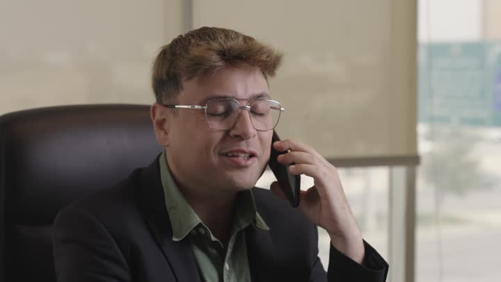 اجراء مكالمات هاتفية خاصة بالعمل , رجل اجنبي يرتدي البذلة الرسمية و نظارات يجلس على المقعد الجلدي داخل المكتب , تعابير الاحترافية و السعادة , تقريب المسافات و توفير الوقت باستخدام الاجهزة التقنية الحديثة