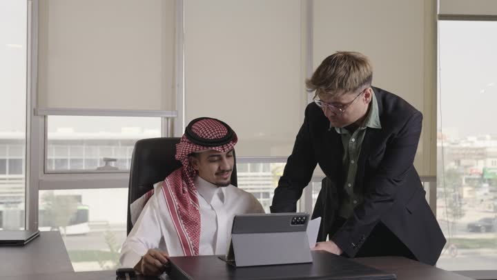 متابعة المعلومات المسجلة على الجهاز اللوحي , رجل عربي خليجي سعودي يرتدي الثوب الابيض و الشماغ الملون يراجع بعض الافكار الخاصة بالمشروع , رجل اجنبي يرتدي البذلة الرسمية و النظارات يؤكد على المعلومات الموجودة على الجهاز
