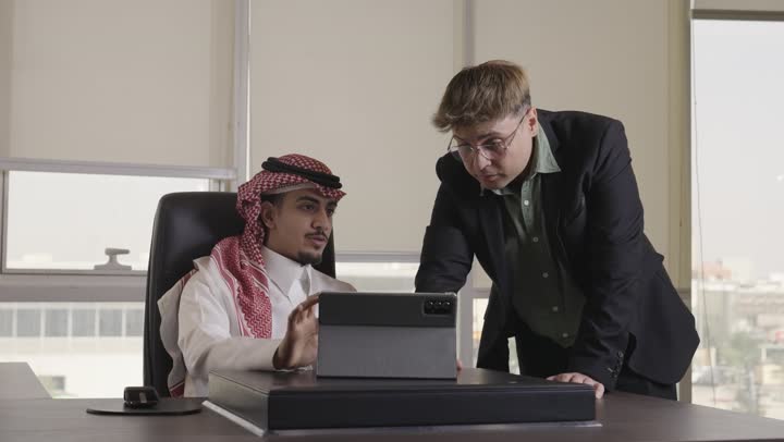 الاشارة باصبع السبابة الى شئ ما في شاشة الجهاز اللوحي , رجل عربي خليجي سعودي يرتدي الثوب الابيض و الشماغ الاحمر يوضح بعض النقاط ,الجلوس داخل مكتب الشركة , رجل اجنبي يرتدي البذلة الرسمية و نظارات يستمع بانصات الى التوضيح