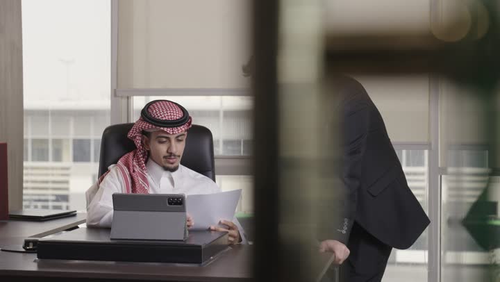 الاستماع بانصات الى توجيهات مدير العمل , رجل اجنبي يرتدي البذلة الرسمية و النظارات ينظر الى الاوراق في يد المدير , رجل عربي خليجي سعودي يرتدي الثوب الابيض و الشماغ الاحمر يحمل بعض التقارير الخاصة بالمشروعات
