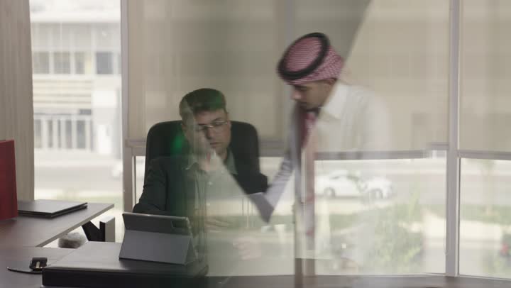 A Saudi Arabian Gulf man wearing a white thobe and a red ghutrah is preparing the important project documents, while a foreign expert wearing glasses and a formal suit is comparing the information on paper with what is recorded on the tablet, displaying expressions of seriousness and professionalism.