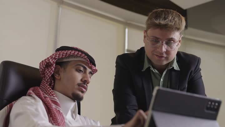 تبادل الافكار و الاراء المهمة لتنفيذ المشروع بالشكل الافضل , رجل عربي خليجي سعودي يرتدي الثوب الابيض و الشماغ الاسود يجلس على المكتب يتحدث الى زميل العمل , رجل اجنبي يرتدي البدلة الرسمية يوضح بعض النقاط الخاصة بالعمل