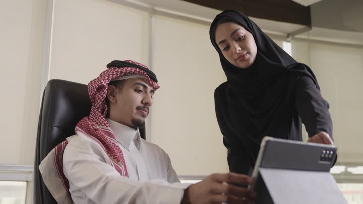 رجل عربي خليجي سعودي يرتدي الشماغ الملون و الثوب الابيض يحرك الراس علامة على الموافقة و الاعجاب بطريقة اداء الاعمال , امرأة عربية خليجية سعودية محجبة ترتدي العباءة السوداء تبين بعض النقاط المهمة في التقارير , الاشارة باصبع السبابة الي جزء ما في الاوراق