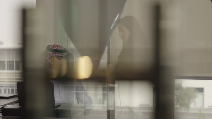 تقديم الاوراق الخاصة بالمشروع للمدير , أمراة عربية خليجية سعودية محجبة ترتدي العباءة السوداء تعطي المدير الملفات المهمة تخص المشروع , رجل عربي خليجي سعودي يرتدي الثوب الابيض و الشماغ الملون يجلس امام المكتب داخل مقر الشركةو استخدام الاجهزة التقنية الحديثة لاتمام العمل بطريقة سلسة
