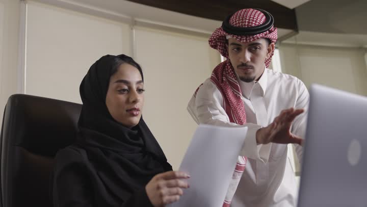 مهندس عربي خليجي سعودي يرتدي الثوب الابيض و الشماغ الملون يوضح العمل المطلوب للزميلة في الفريق , مرأة عربية خليجية سعودية محجبة ترتدي العباءة السوداء تحمل الاوراق المطلوب تدوينها على الحاسب المتنقل