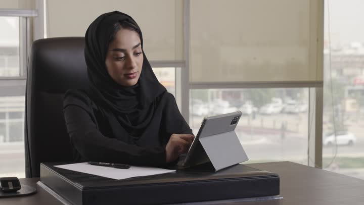 تسجيل التقارير الخاصة بالمشاريع الهندسية , امرأة عربية خليجية سعودية محجبة ترتدي العباءة السوداء تقوم بنقل الملاحظات من الجهاز اللوحي التقني , الجلوس امام المكتب الخشبي