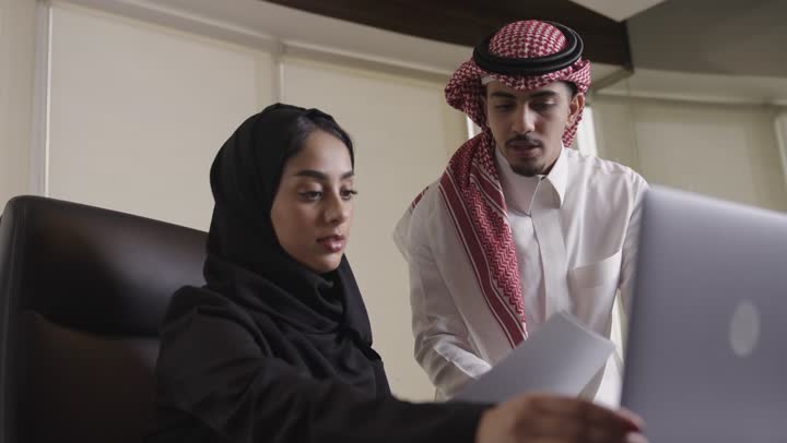 مقارنة المعلومات الورقية بالموجودة على الحاسب المتنقل , رجل عربي خليجي سعودي يرتدي الثوب الابيض و الشماغ الملون يتابع بدقة المعلومات المدونة بالورق , امراة عربية خليجية سعودية محجبة ترتدي العباءة السوداء تحمل الاوراق الخاصة بالمشروعات , مفهوم العمل الجماعي