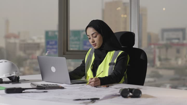مرأة عربية خليجية سعودية ترتدي العباءة السوداء تعمل على المشروع الهندسي , استخدام الاجهزة التقنية الحديثة في انجاز المطلوب بدقة , ايماءات التركيز و الاحترافية العالية