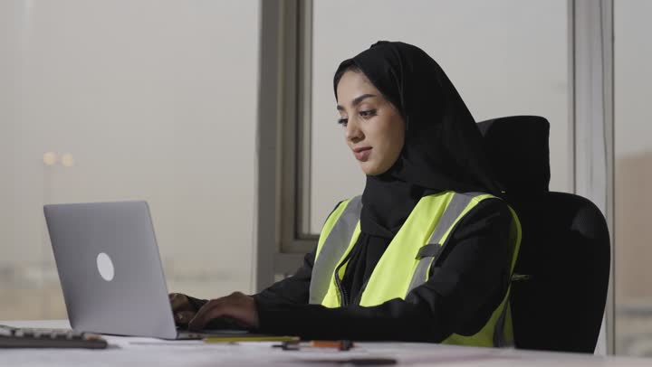 الدقة و الاحترافية في تنفيذ المشاريع الهندسية , مهندسة عربية خليجية سعودية محجبة ترتدي العباءة السوداء تسجل الملاحظات على الحاسب المتنقل , الجلوس داخل مبنى الشركة