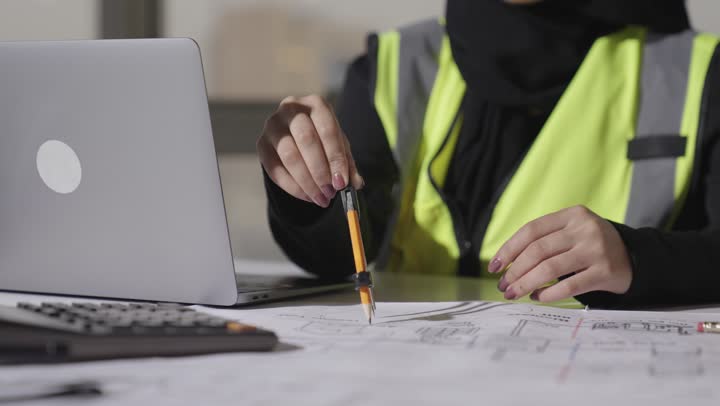 تصوير مقرب لمهندسة عربية خليجية سعودية محجبة ترتدي العباءة السوداء تستخدم الادوات الهندسية في رسم المخططات الهندسية , العمل بدقة و تركيز