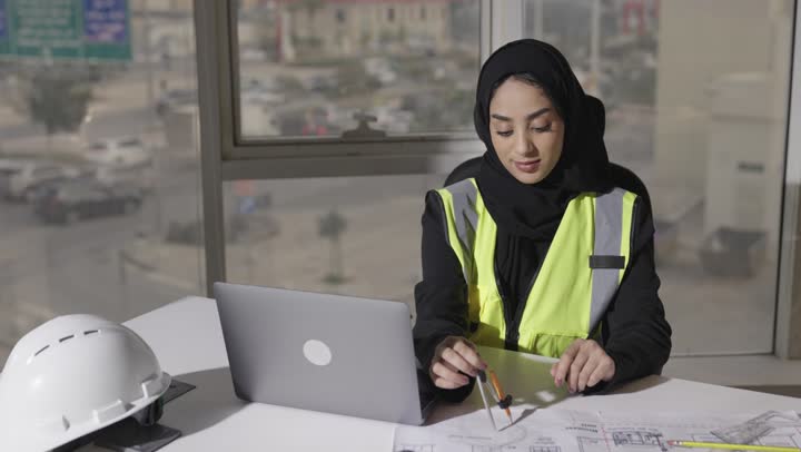 The use of engineering tools in drawing architectural plans, a veiled Arab Gulf Saudi engineer wearing a black abaya and a protective jacket is performing the required work with high professionalism using a portable computer.