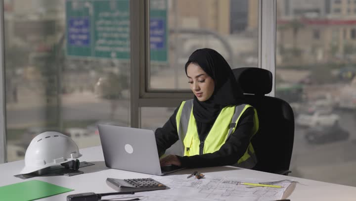كتابة التقارير اليومية و الرسومات الهندسية الخاصة بالمشارع المعمارية باستخدام الاجهزة التقنية الحديثة , امرأة عربية خليجية سعودية محجبة ترتدي عباءة سوداء تجلس امام المكتب تتابع عملها اليومي , مفهوم توفير الجهد و تنفيذ العمل باحترافية