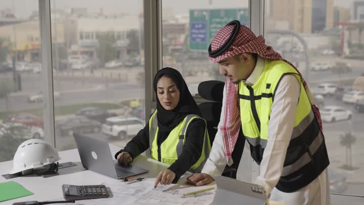 Comparison of engineering plans on drawn papers and those available on the portable computer. An Arab Gulf Saudi engineer wearing a black abaya presents the design on the modern technical device. An Arab Gulf Saudi engineer wearing a white thobe and a colored ghutrah presents his personal perspective on the project, the concept of teamwork.