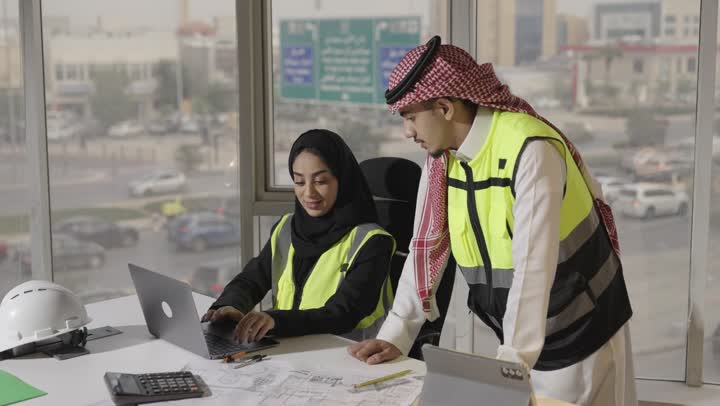 مهندسة معمارية عربية سعودية خليجية محجبة ترتدي عباءة سوداء تتابع الاعمال الهندسية اليومية على جهاز الحاسب المتنقل , مهندس عربي خليجي سعودي يرتدي الشماغ الاحمر و الثوب الابيض يرسم خط طولي باصبع السبابة على المخطط الهندسي , تبادل الاراء و المعلومات بحرفية ودقة