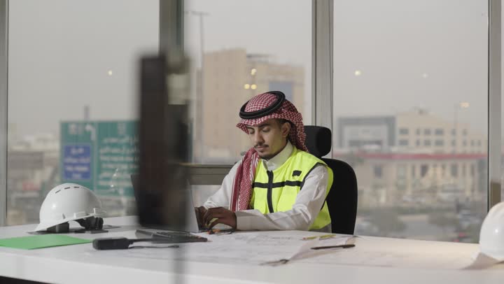 مشاركة الرأي و العمل الجماعي في بيئة عمل صحية , مهندسة عربية خليجية سعودية ترتدي العباءة السوداء تحمل الجهاز اللوحي , مهندس عرب خليجي سعودي يرتدي الثوب الابيض و الشماغ الملون يجلس امام المكتب ويقدم الاستشارات الهندسية للزميلة