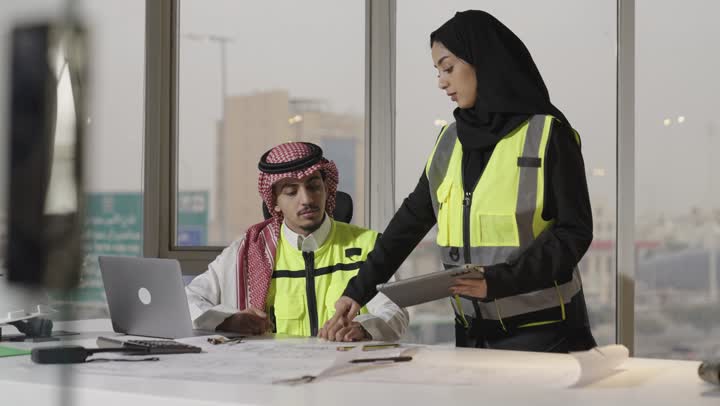 الاشارة باصبع السبابة الى جزء ما في المخططات الهندسية , رجل عربي خليجي سعودي يرتدي السترة الواقية و الثوب الابيض يتابع الاعمال اليومية باستخدام الاجهزة التقنية الحديثة , مهندسة عربية خليجية سعودية ترتدي العباءة السوداء تعرض لزميل العمل المعلومات حول المشروع القائم