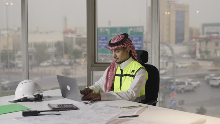 العمل الهندسي بسهولة باستخدام الاجهزة التقنية الحديثة , رجل عربي خليجي سعودي يرتدي الثوب الابيض و الشماغ الملون يجلس امام طاولة العمل , مهندسة عربية خليجية سعودية محجبة ترتدي العباءة السوداء و السترة الواقية تشارك زميل العمل الرأي في المشروع قيد التنفيذ