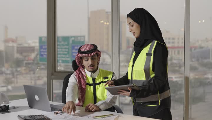 Exchange of opinions and observations about the ongoing project, a Saudi Gulf Arab engineer wearing a white thobe and a colored ghutrah points with his fingers to the engineering plans on the desk, a Saudi Gulf Arab female engineer wearing a black abaya and a hijab holds a tablet and shares information with a colleague.