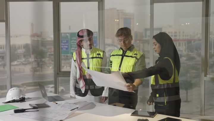 رجل عربي خليجي سعودي يرتدي الشماغ الملون و الثوب الابيض يقف بجانب زملاء العمل , مهندس اجنبي يرتدي الزي الكاجوال يحمل المخطط الهندسي يستمع الى اراء باقى المهندسين , امراة عربية خليجية سعودية محجبة ترتدي العباءة السوداء تقدم الملاحظات المهمة الخاصة بالمشروع الهندسي