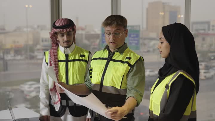 محادثة بين اعضاء فريق العمل حول التخطيط العمراني المرسوم , رجل اجنبي يرتدي الزي الكاجوال يحمل المخطط الهندسي , امرأة عربية خليجية سعودية محجبة ترتدي عباءة سوداء تشير باصبع السبابة الى شئ ما في المخططات الهندسية , مهندس عربي خليجي سعودي يرتدي السترة الواقية و الثوب الابيض يستمع بانصات , مفهوم العمل الجماعي