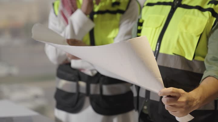 A Saudi Arabian Gulf man wearing a white thobe and a protective vest is closely following an explanation of the engineering plans, a foreign man in casual attire is holding the engineering plan for the project, and an Arab Gulf woman is pointing out some notes in a certain part, close-up shot.