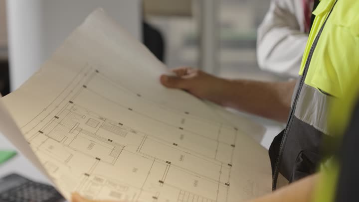 رجل اجنبي يرتدي السترة الواقية العاكسة للضوء يحمل المخطط الهندسي , امرأة عربية خليجية سعودية ترتدي العباءة السوداء تشير باصبع السبابة الي شئ ما في الرسومات الهندسية , مهندس عربي خليجي سعودي يرتدي الثوب الابيض يقدم الاستشارات حول المخطط المعماري