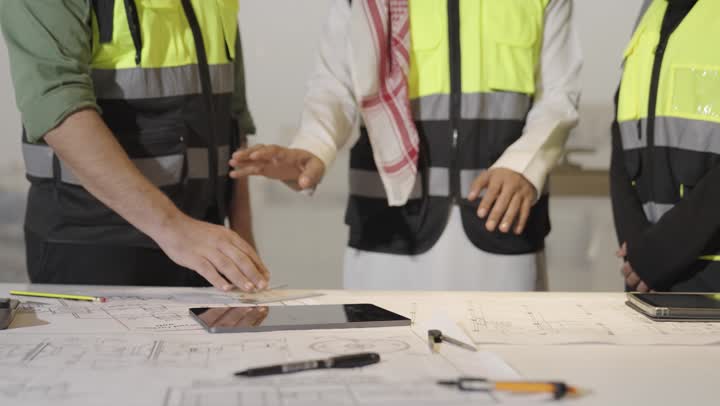 الاشارة الي جزء بعيد في المخطط الهندسي , رجل عربي خليجي سعودي يرتدي الثوب الابيض و الشماغ الملون يشير الى الرسومات , مهندسة عربية خليجية سعودية محجبة ترتدي عباءة سوداء تقف مع فريق العمل , رجل اجنبي يرتدي الزي الكاجوال يحمل القلم الرصاص لاضافة التعديلات على التخطيط