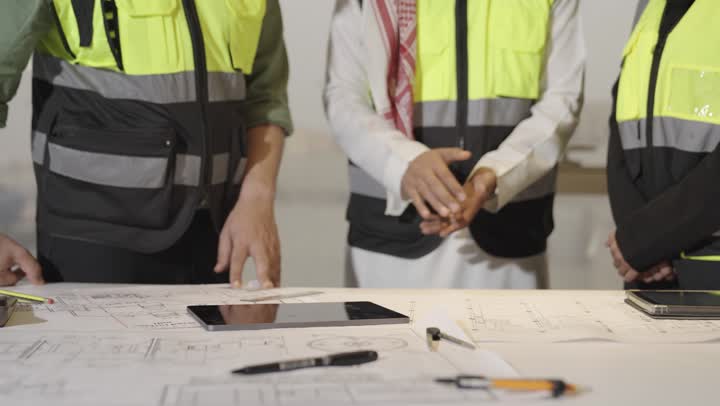 مدير الفريق العربي الخليجي السعودي يرتدي لسترة الواقية يرفع اصبع الابهام علامة على الموافقة على اقتراحات الزملاء , مهندسة عربية خليجية سعودية ترتدي العباءة السوداء تقدم المقترحات المهمة لمساعدة الفريق , رجل اجنبي يرتدي الزي الكاجوال يشير الي الرسم الهندسي , مفهوم العمل الجماعي , الوقوف حول المكتب